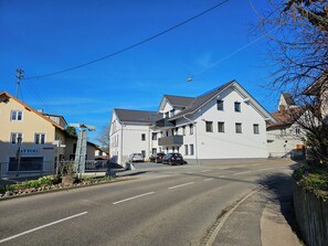 A street view with a building labeled "Tattoo" on the left, a white building with a balcony in the center, and a clear blue sky.