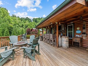 Una terraza de madera con sillas Adirondack, un hogar de piedra y una zona de bar con taburetes de madera.