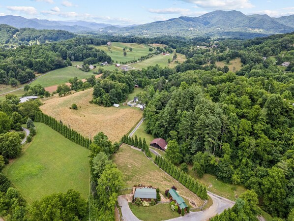 Vista aérea de un paisaje rural con un extenso campo verde, un pequeño conjunto de edificios y densas áreas boscosas.