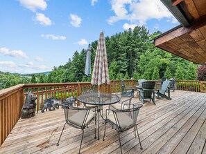 Una terraza de madera con mobiliario exterior, incluyendo sillas y una mesa, y vistas a un paisaje boscoso.