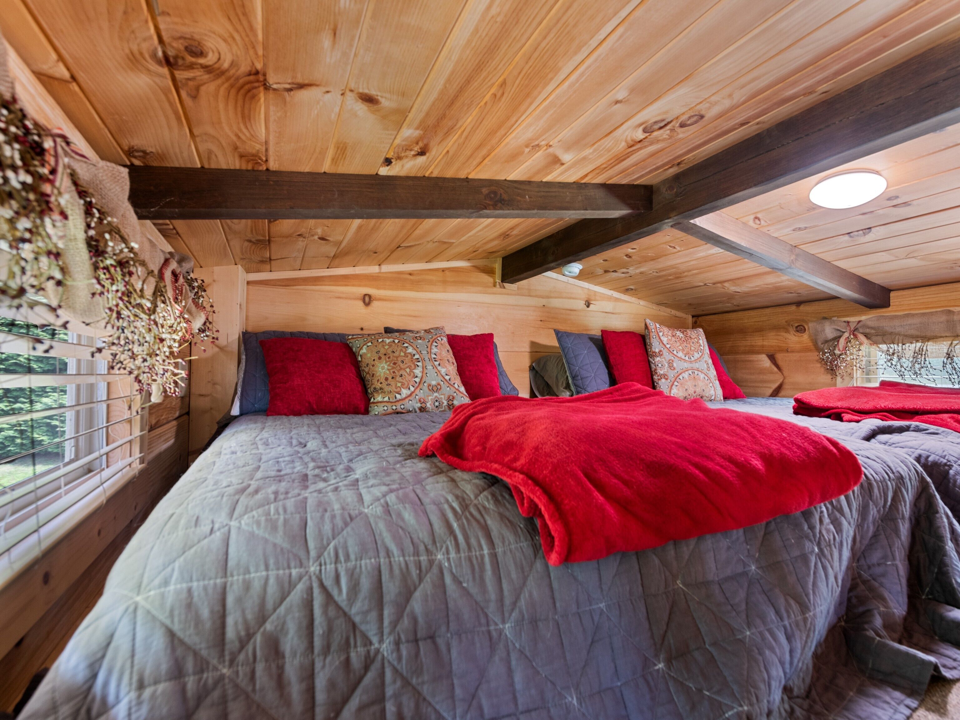 Dormitorio de cabaña de madera con una cama grande, varios almohadones y ventana con vista a la vegetación.