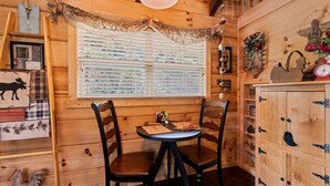 Interior de una cabaña de madera con una mesa redonda, dos sillas, una ventana con persianas, un armario de madera y varios adornos.