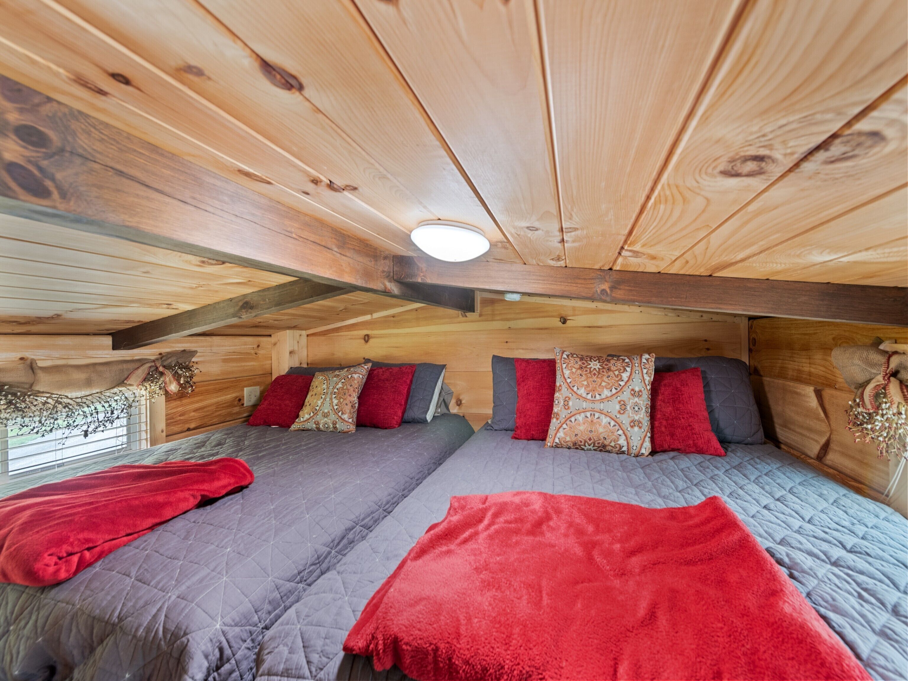 A cozy cabin room with wooden walls and ceiling, two beds with red and grey bedding, and a window with a view of the forest.