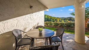 An outdoor patio with a round table and four wicker chairs, a potted plant, and a view of a landscaped garden and hills.
