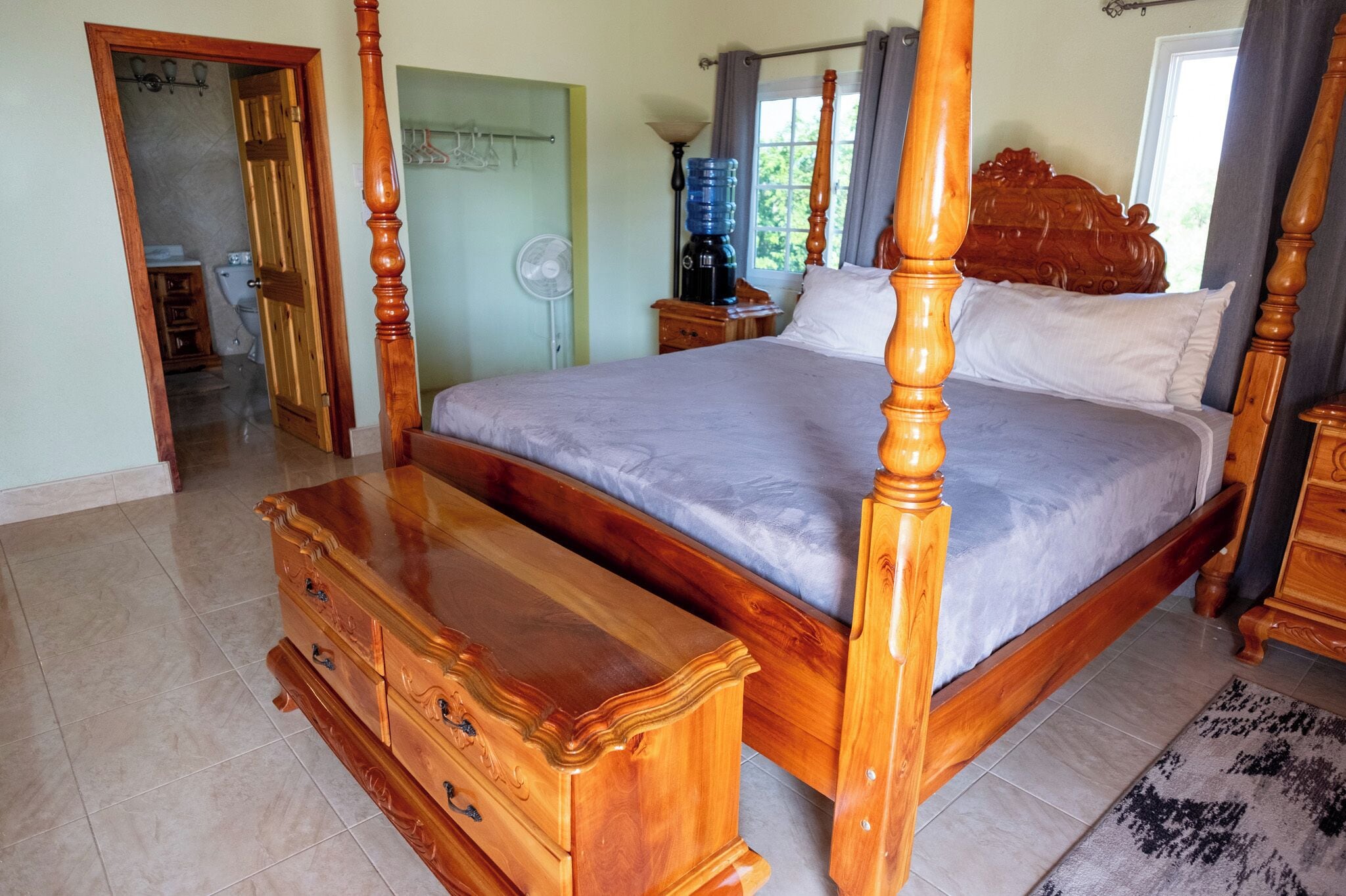 Una cama con dosel, un cabecero de madera, un aparador de madera con cajones y piso de baldosas.