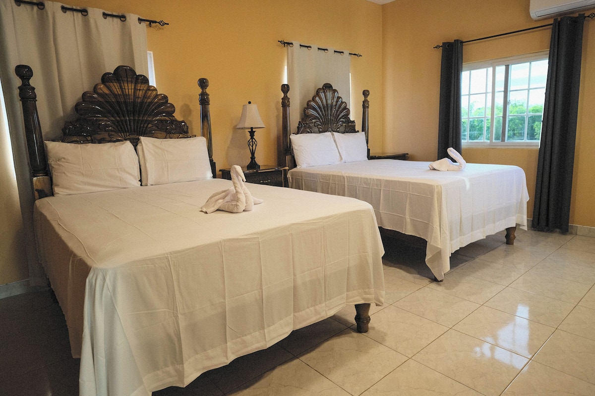 A hotel room with two beds, a nightstand, a lamp, and a window with curtains.
