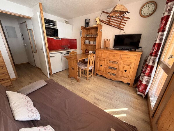 A compact room with a bed, a wooden dining table with chairs, a kitchenette with a microwave, and a TV.