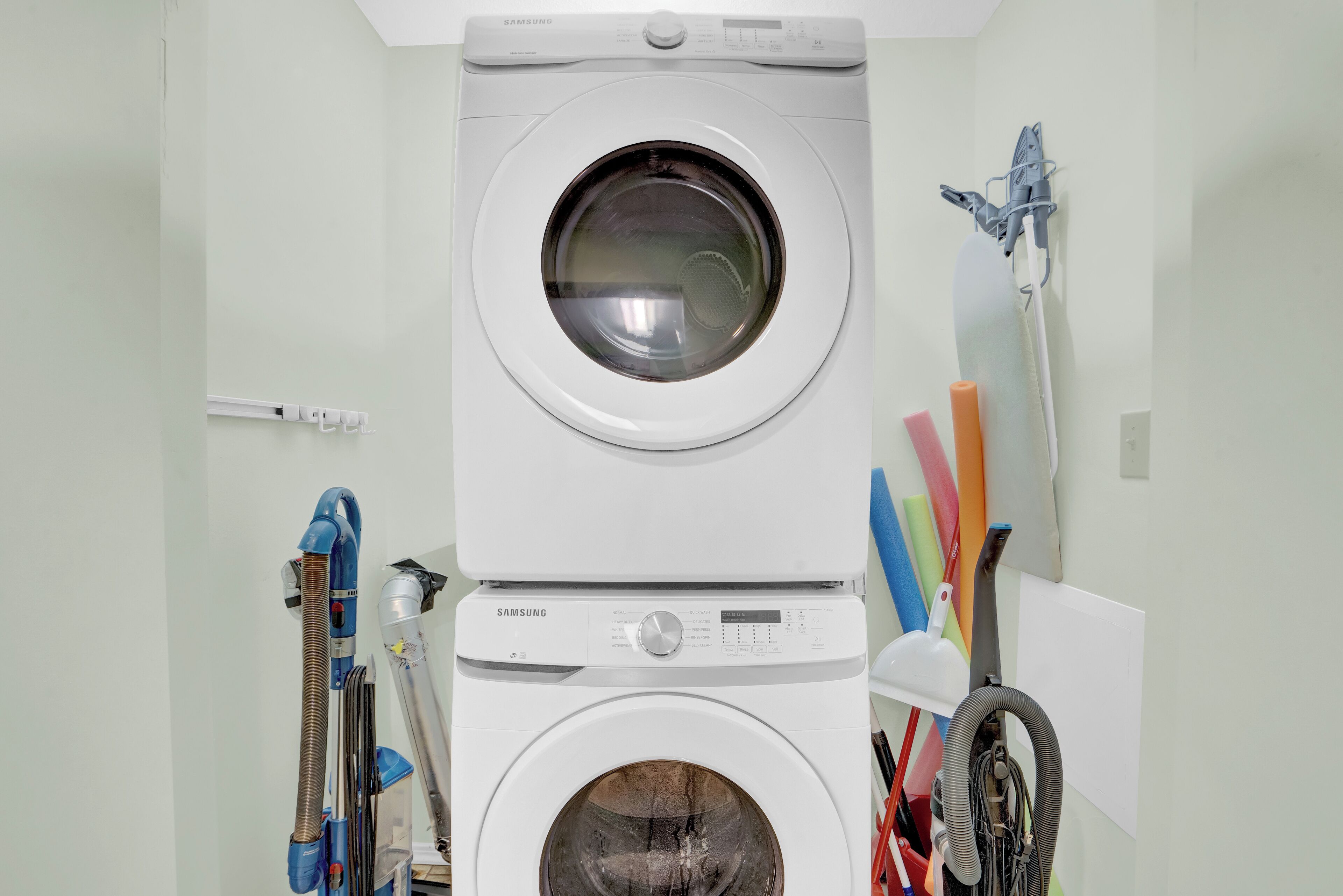 A stacked washer and dryer unit in a laundry room with various cleaning tools hanging on the wall.