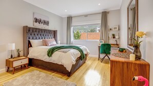 Un dormitorio con cama y cabecera capitonada, una mesita de noche con lámpara, un escritorio de madera con silla, un ventanal con cortinas y un espejo en la pared.