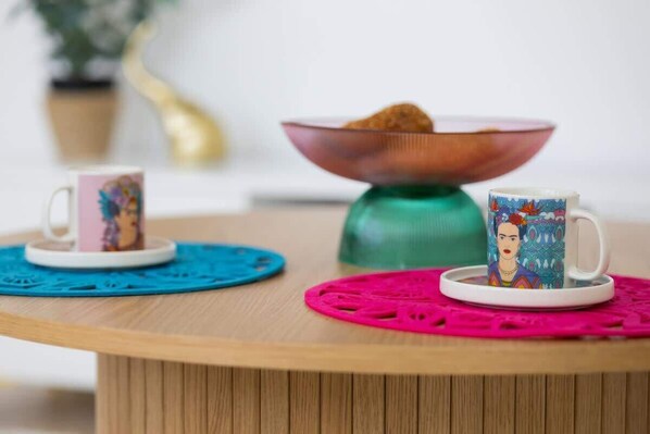 A round wooden table with two ceramic cups on matching saucers, a decorative bowl, and a potted plant in the background.