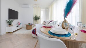 Un salon moderne avec un coin repas, comprenant une table ronde en bois, des chaises blanches, un téléviseur à écran plat fixé au mur et une grande fenêtre ornée de rideaux légers.