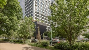 Un immeuble moderne de grande hauteur, doté d’une façade en verre, entouré d’une végétation luxuriante et de voitures garées.