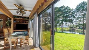 A modern living room with a glass dining table, wooden chairs, a ceiling fan, and a large sliding glass door leading to a grassy outdoor area with trees. - Sandy Beaches *Heated Pool* Pet Friendly (Destin)