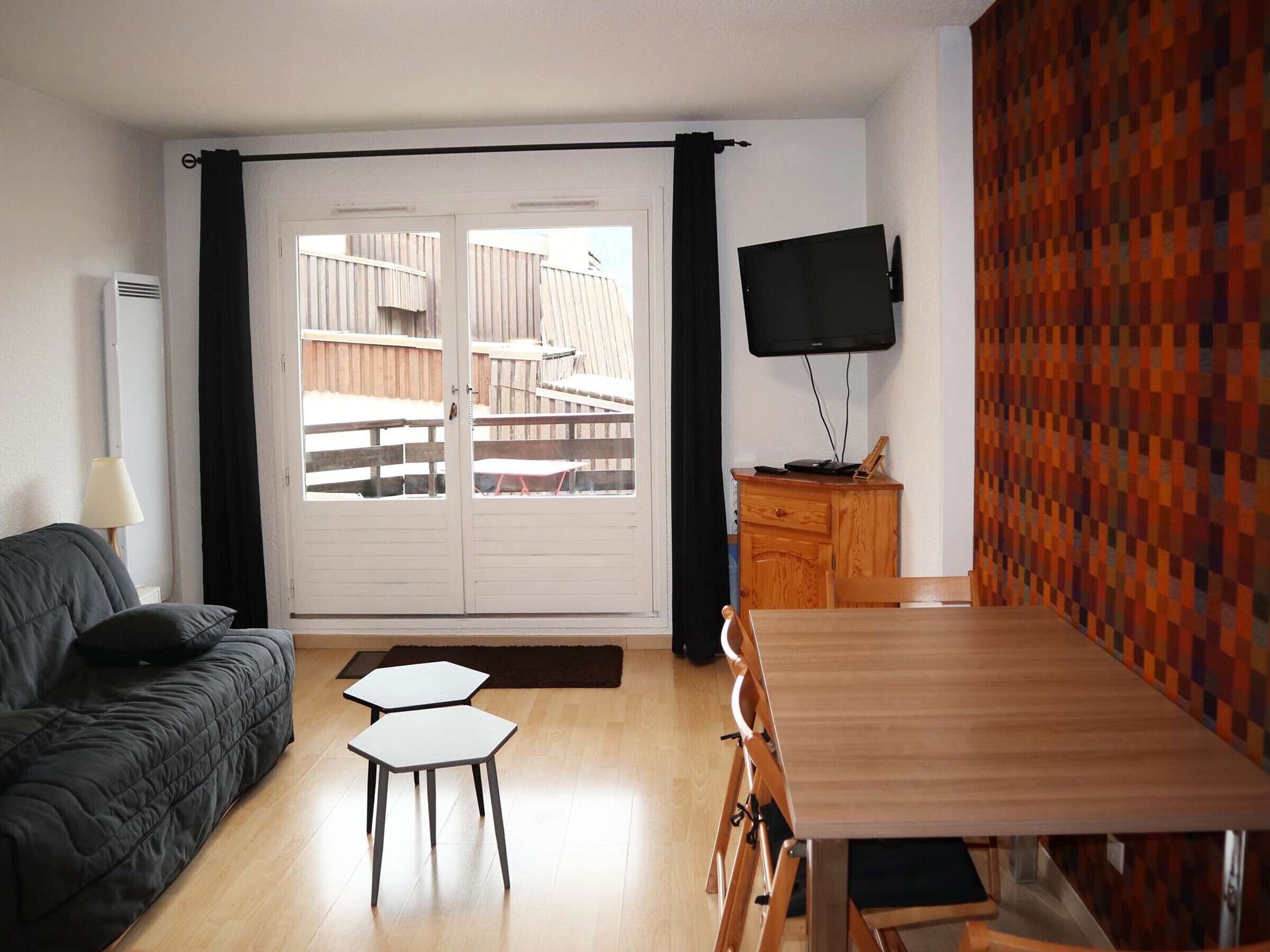 Une pièce comprenant un canapé, une table à manger avec des chaises, un téléviseur fixé au mur et une vue accessible par une porte coulissante.