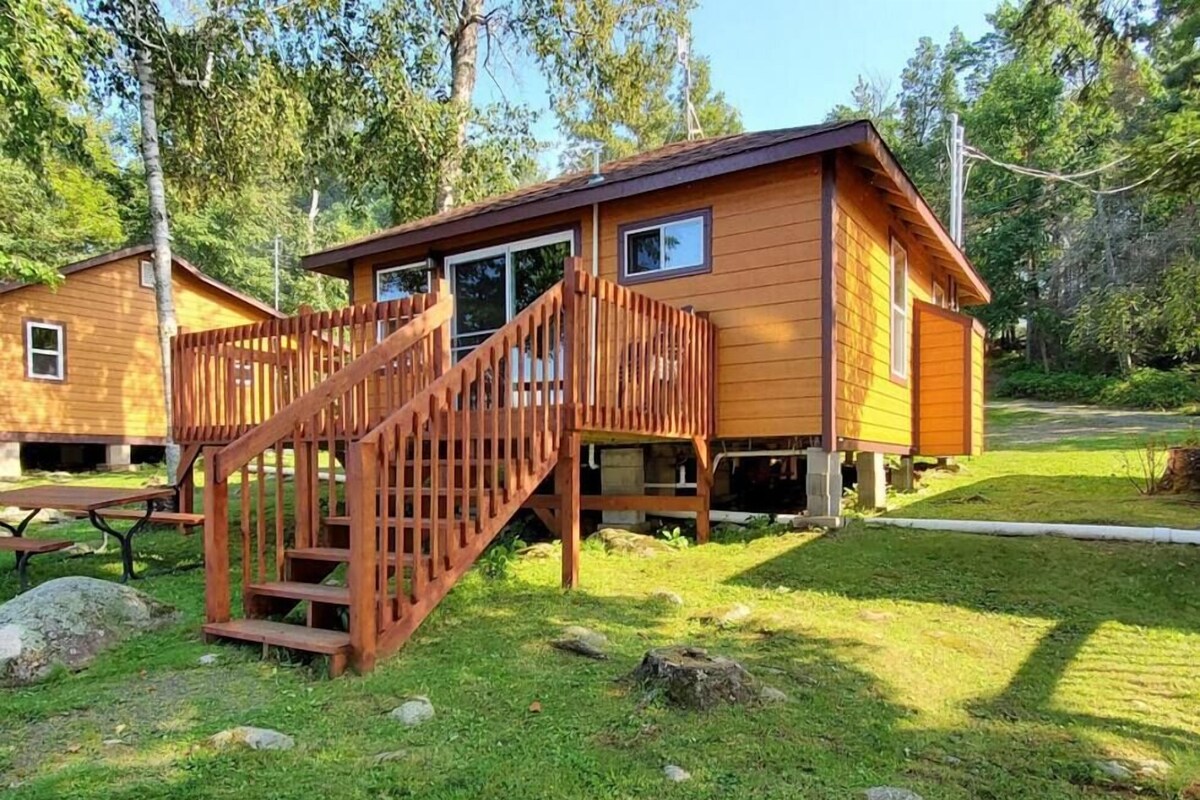 Une cabane en bois avec un escalier menant à une terrasse, entourée d’une pelouse et d’arbres.