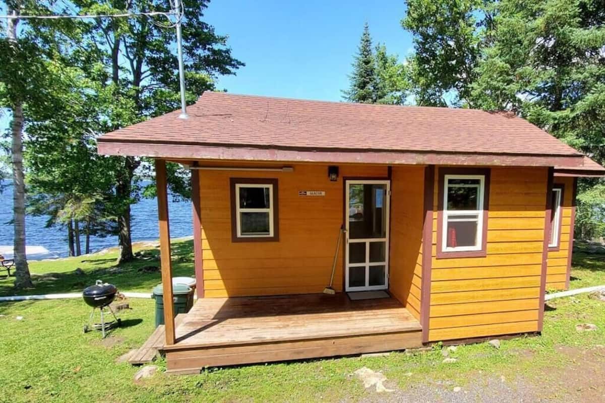 Une petite cabane en bois dotée d’une véranda, d’une porte et de fenêtres, située près d’une étendue d’eau.