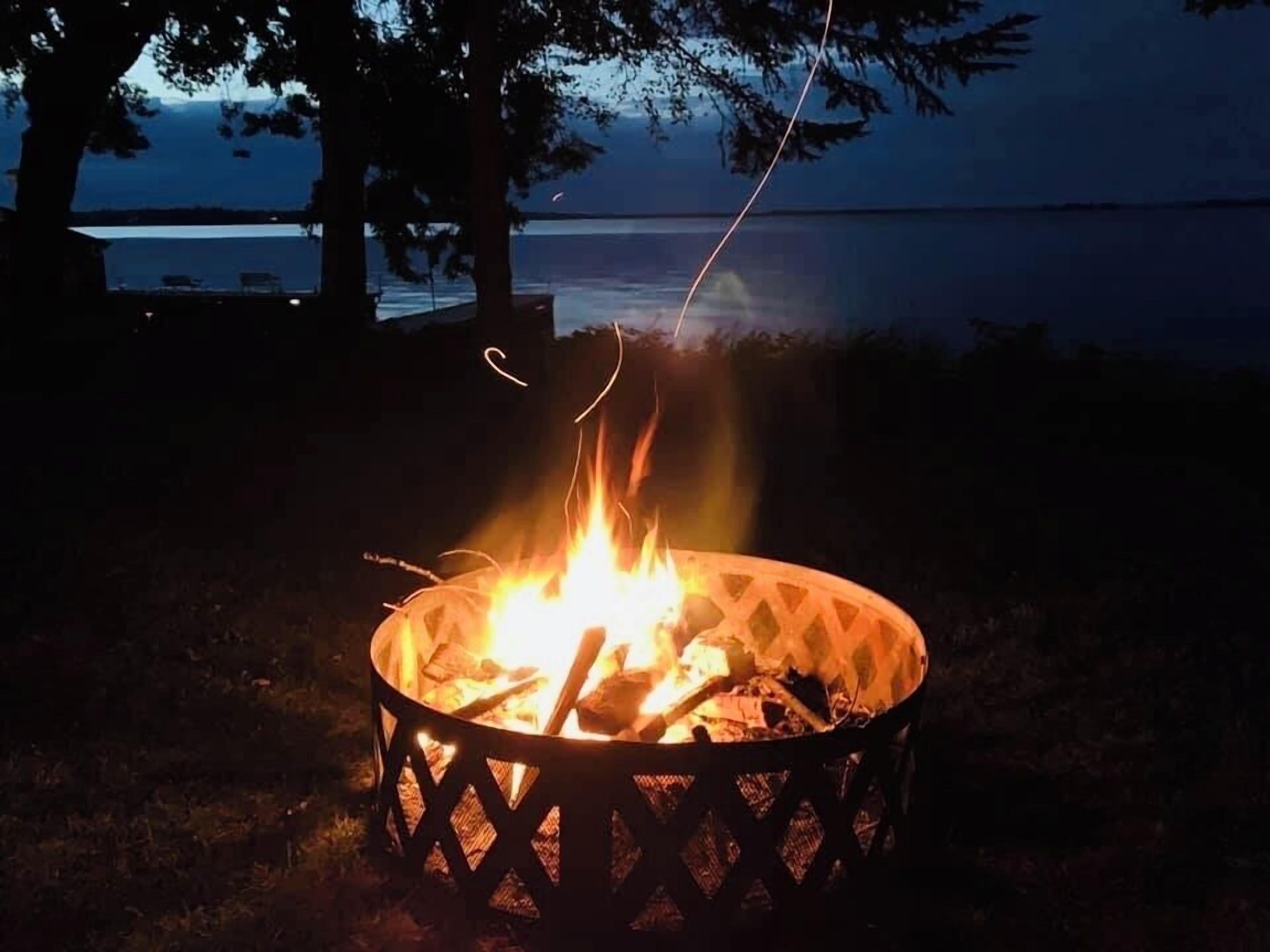A campfire at dusk with trees and a lake in the background.