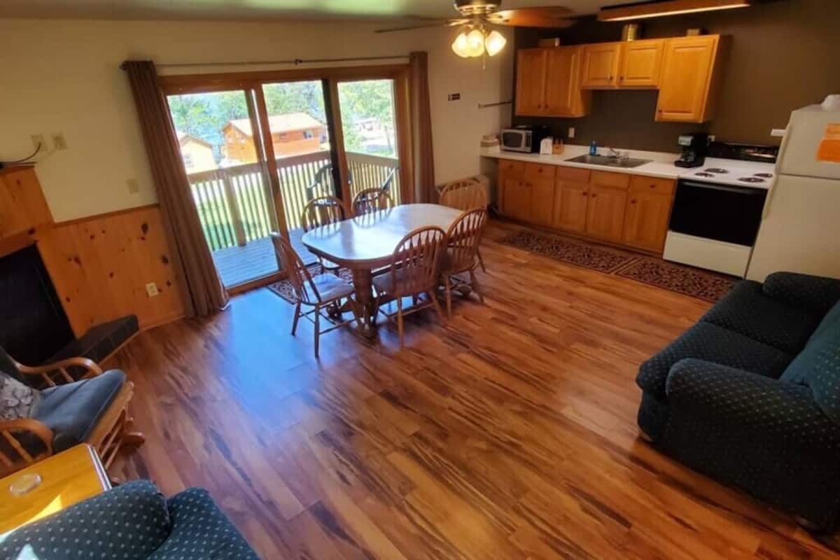 Un salon chaleureux avec un plancher en bois, une table à manger avec des chaises, un canapé et un coin cuisine doté de meubles et d’appareils électroménagers en bois.