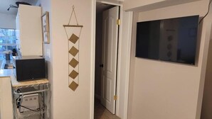 Un pasillo con una puerta que da a una habitación, un televisor montado en la pared y una zona de cocina con un microondas y otros electrodomésticos.