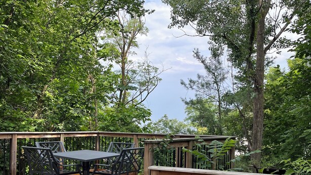 An outdoor deck with wooden flooring and metal furniture, surrounded by lush greenery and trees.