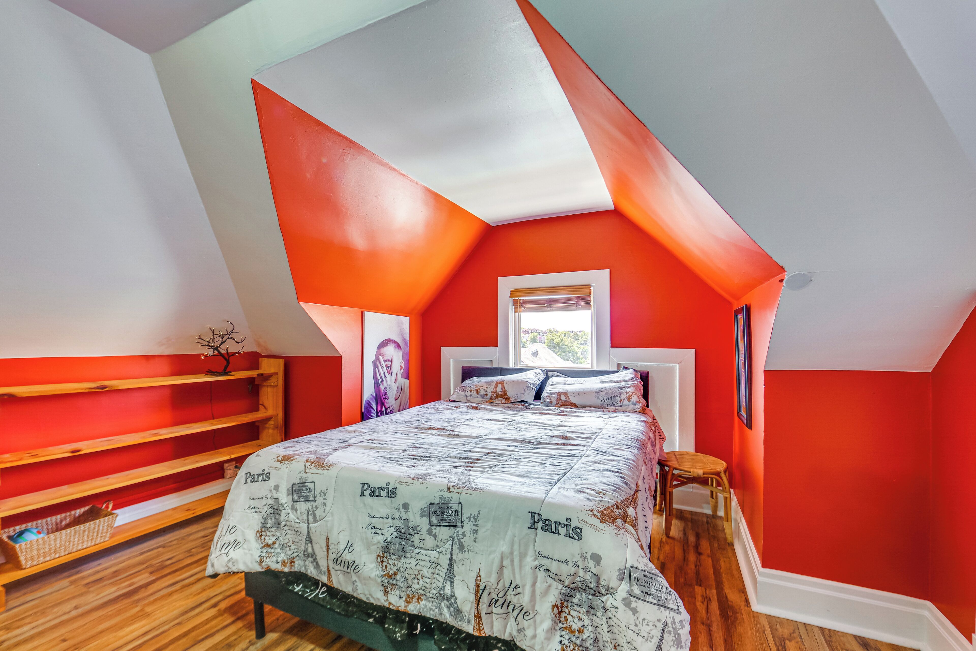 A bedroom with a sloped ceiling, a bed with a Paris-themed bedspread, wooden shelves, and a window with a view.