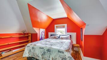 A bedroom with a sloped ceiling, a bed with a Paris-themed bedspread, wooden shelves, and a window with a view.