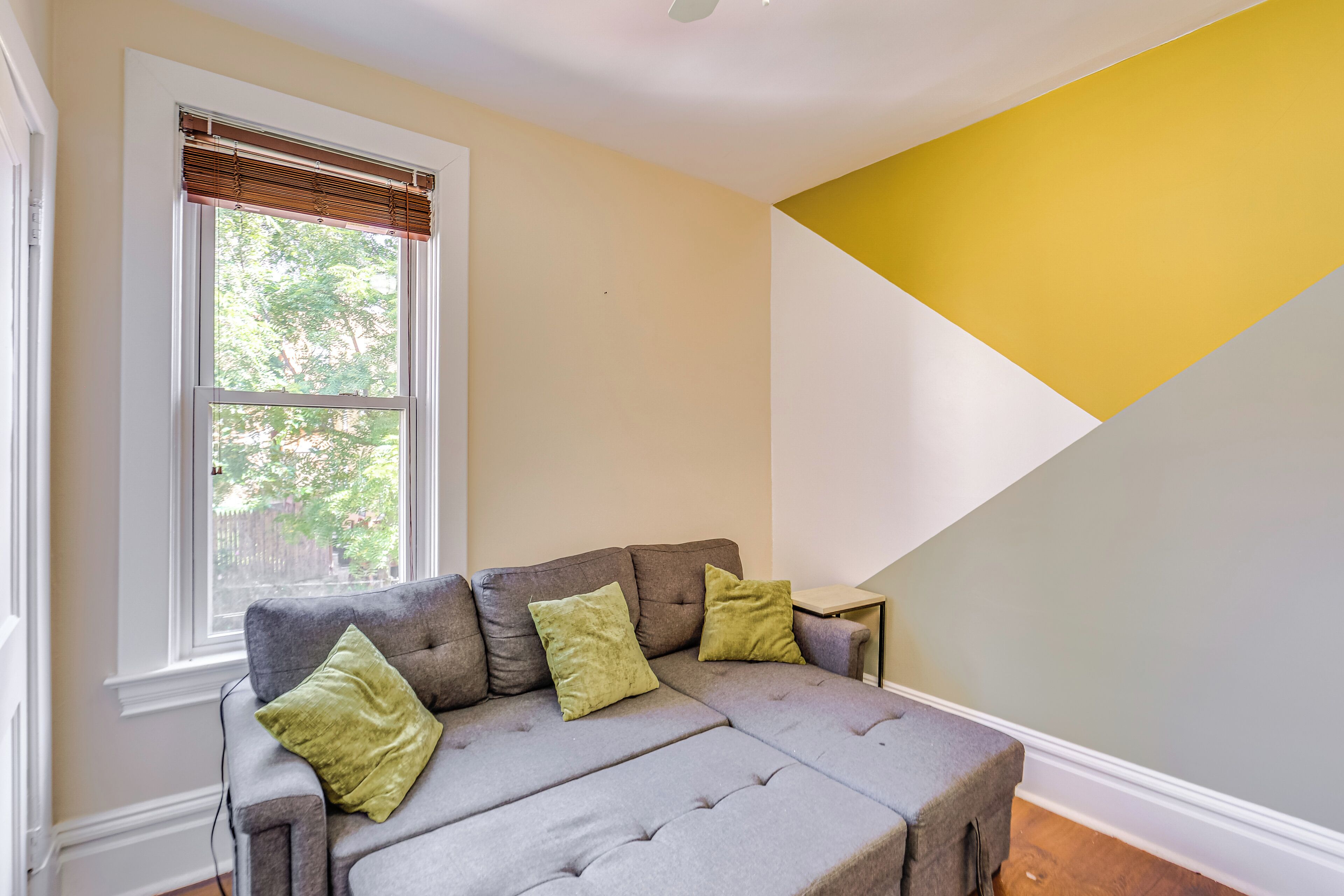 Une pièce avec un canapé gris, deux coussins verts, une fenêtre avec des stores et un mur jaune et blanc.