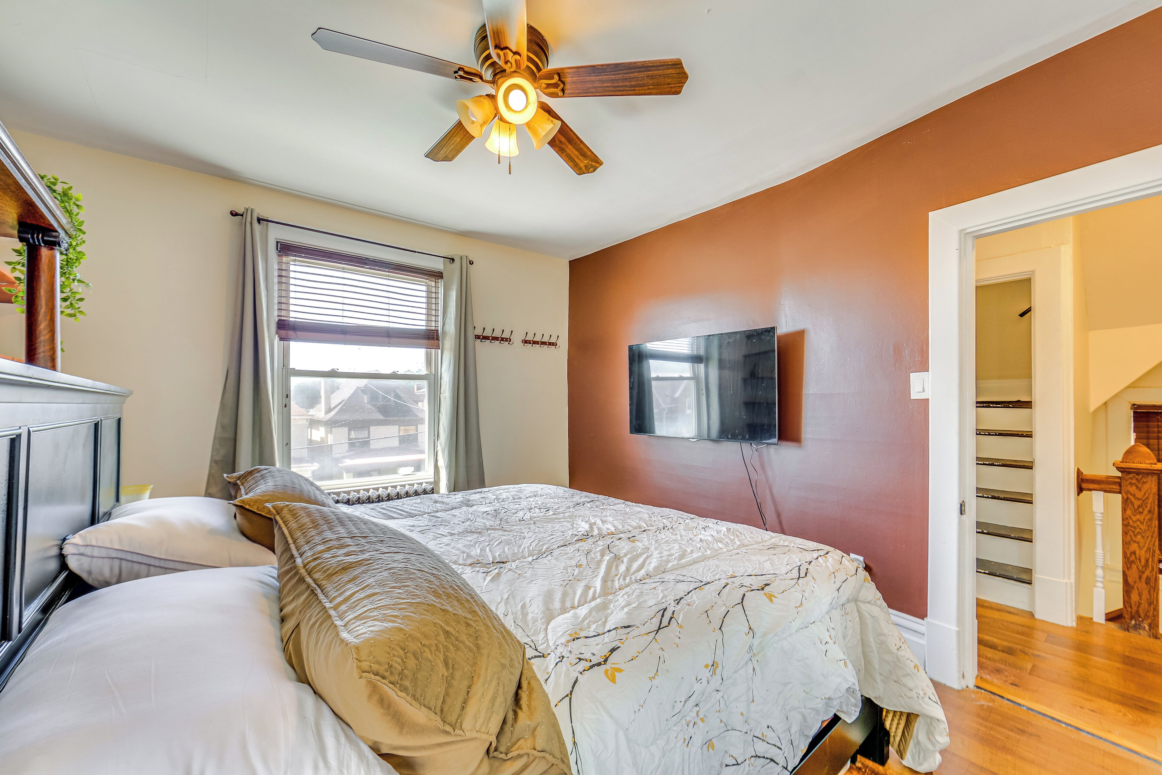 Une chambre à coucher comprenant un lit, un ventilateur de plafond, une fenêtre avec des stores, un téléviseur fixé au mur et un escalier menant à un autre étage.