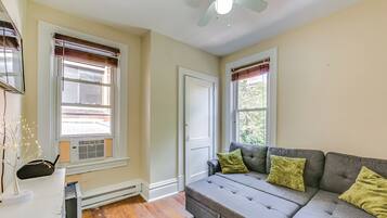 A living room with a grey sofa, two green cushions, a ceiling fan, and a window with blinds.