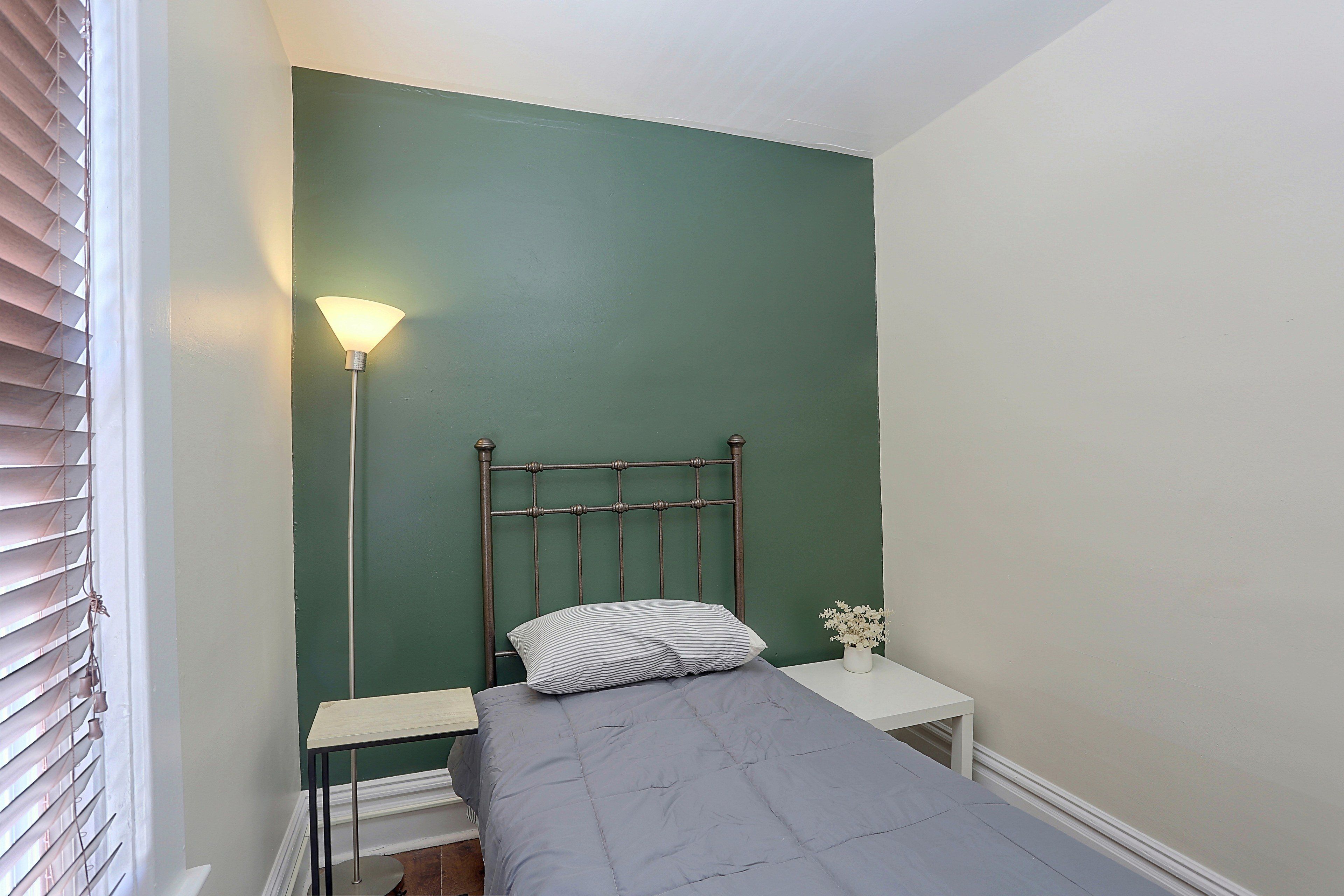 A small, single-bed room with a green wall, a bedside table, a lamp, and a window with blinds.