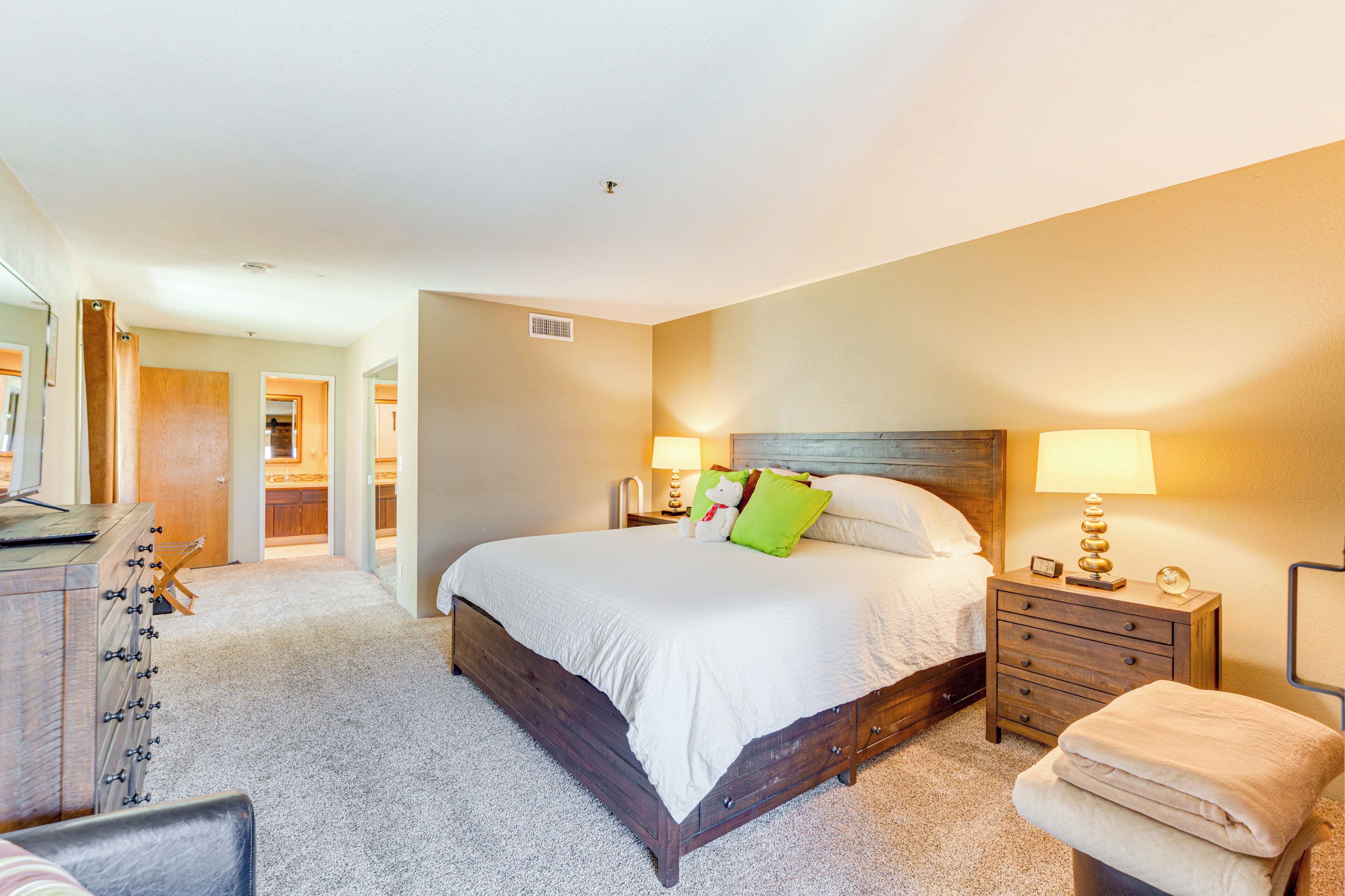 A bedroom with a large bed, a bedside table with a lamp, a dresser, and a chair.