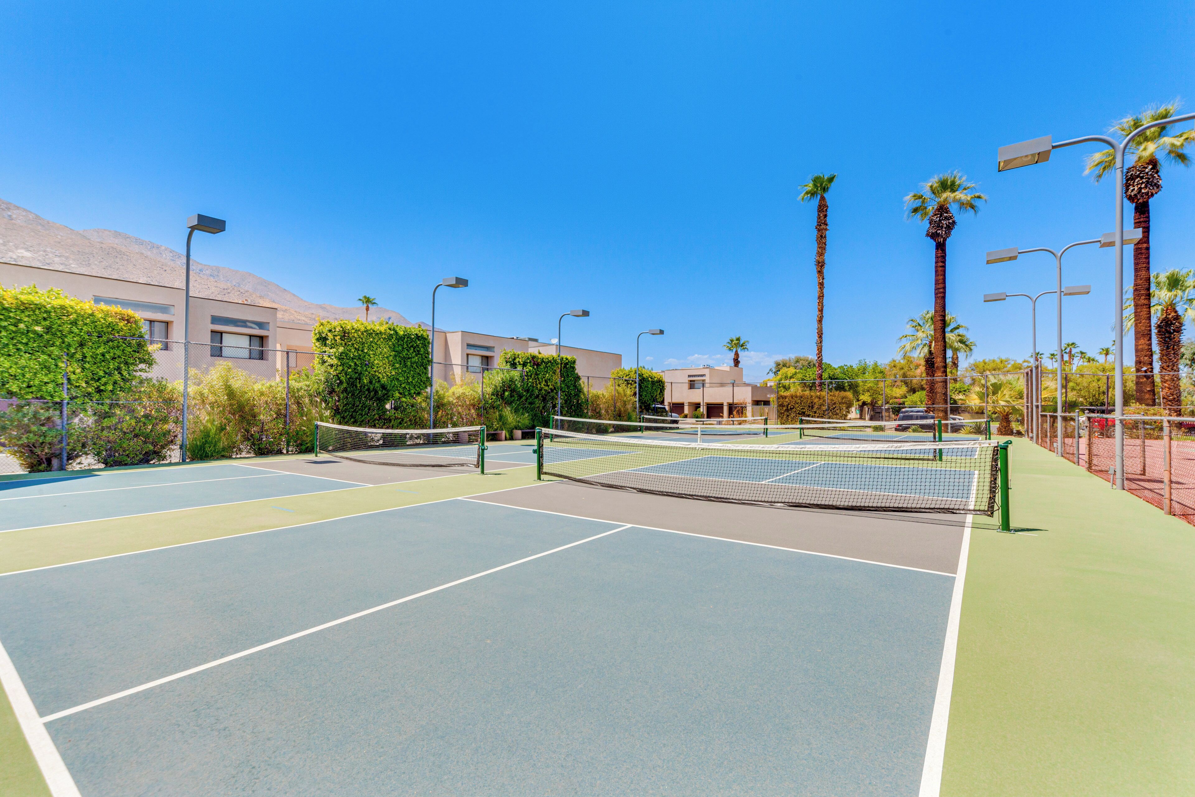 Ein Tennisplatz im Freien mit Netz, umgeben von Palmen und einem Gebäude im Hintergrund.