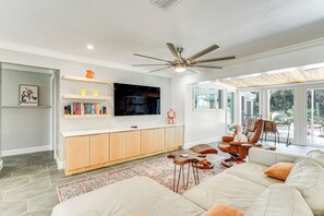A modern living room with a large sofa, a coffee table, and a TV mounted on a wooden cabinet. There is a ceiling fan, a large window, and a view of the outdoors. - Walk to Beach: Cape Coral Home w/ Grill & Patio (Cape Coral)