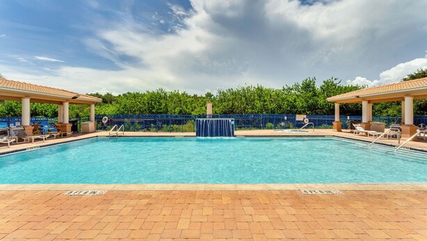 A rectangular swimming pool with a water fountain in the center, surrounded by lounge chairs and umbrellas, with a blue fence and trees in the background. - New Smyrna Beach Condo w/ Balcony & Ocean Walk (New Smyrna Beach)