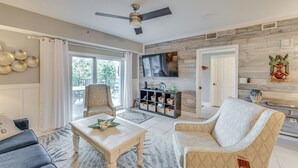 A spacious living room with a ceiling fan, a flat-screen TV mounted on the wall, and a large window with curtains. - New Smyrna Beach Condo w/ Balcony & Ocean Walk (New Smyrna Beach)