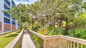 A wooden boardwalk leading to a building with balconies, surrounded by lush greenery and trees. - New Smyrna Beach Condo w/ Balcony & Ocean Walk (New Smyrna Beach)