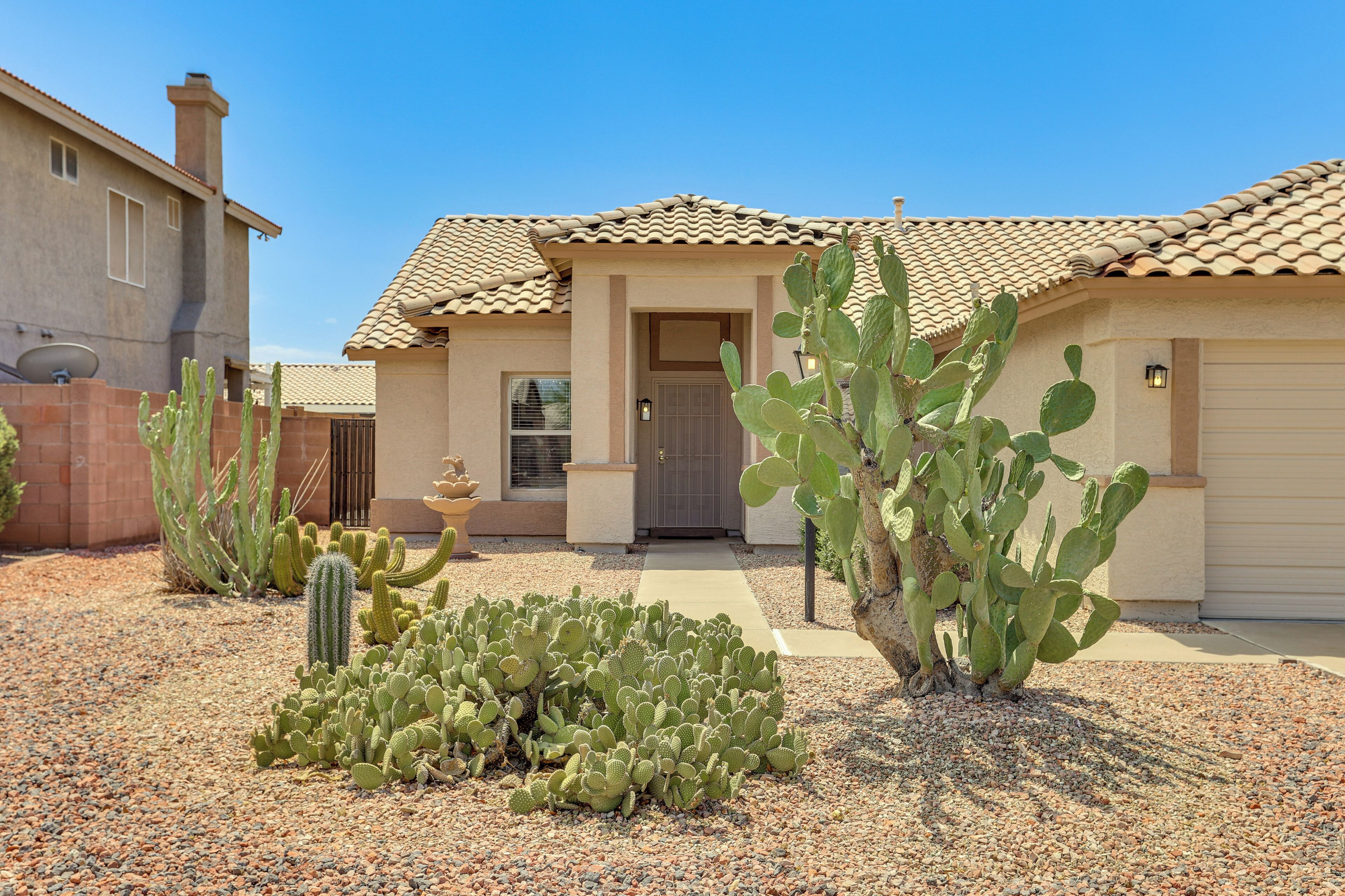 A house with a tiled roof, a front door, and a driveway leading to a garage. There are cacti and a small fountain in the front yard.
