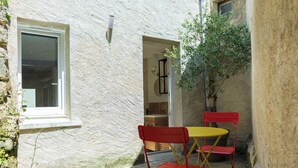 Un petit espace extérieur, avec un mur blanc à la texture particulière, une fenêtre, une petite table dont le plateau est jaune et deux chaises rouges pliantes.