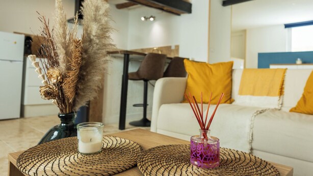 Un salon avec un canapé blanc, des coussins jaunes, une table basse en bois ornée d’une bougie et de plantes séchées, et une cuisine en arrière-plan.
