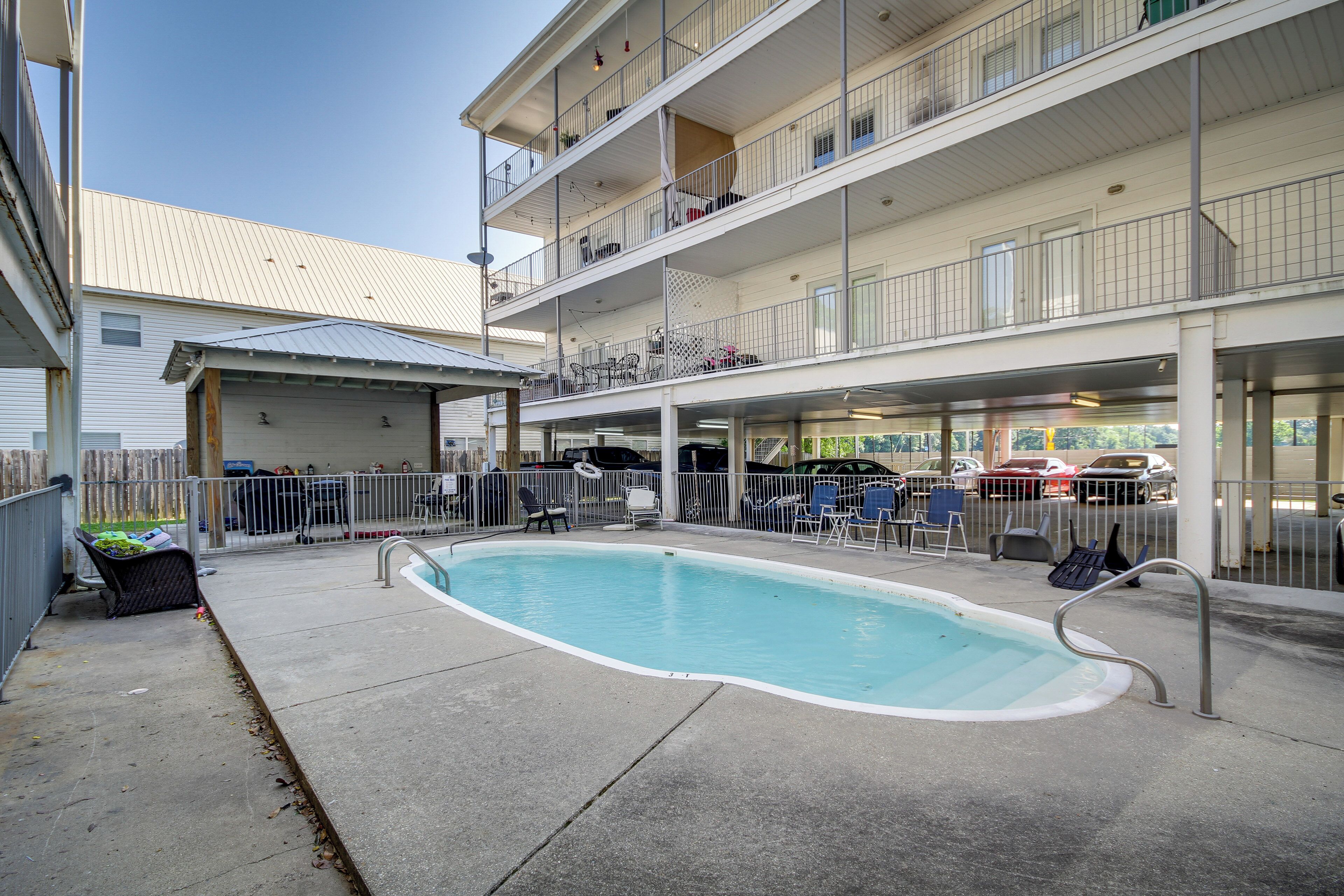 A swimming pool with a concrete deck, surrounded by a metal fence. There are lounge chairs and a covered area with a roof.