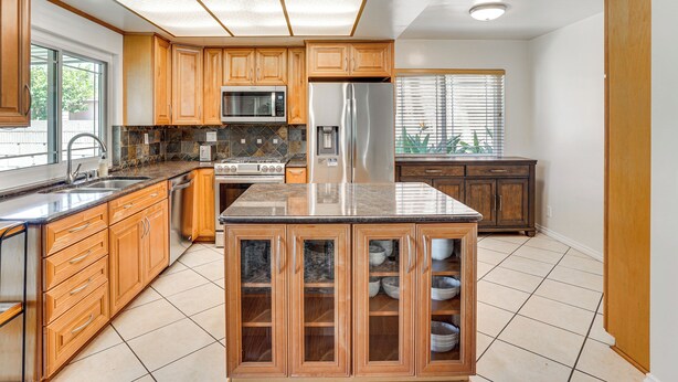 A modern kitchen with wooden cabinets, a central island with glass doors, stainless steel appliances, and a tiled floor. - Huntington Beach Home: Fire Pit, Grill + More! (Huntington Beach)
