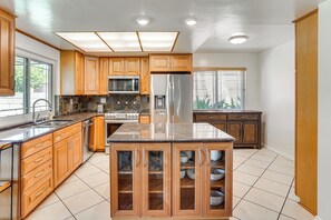 A modern kitchen with wooden cabinets, a central island with glass doors, stainless steel appliances, and a tiled floor. - Huntington Beach Home: Fire Pit, Grill + More! (Huntington Beach)