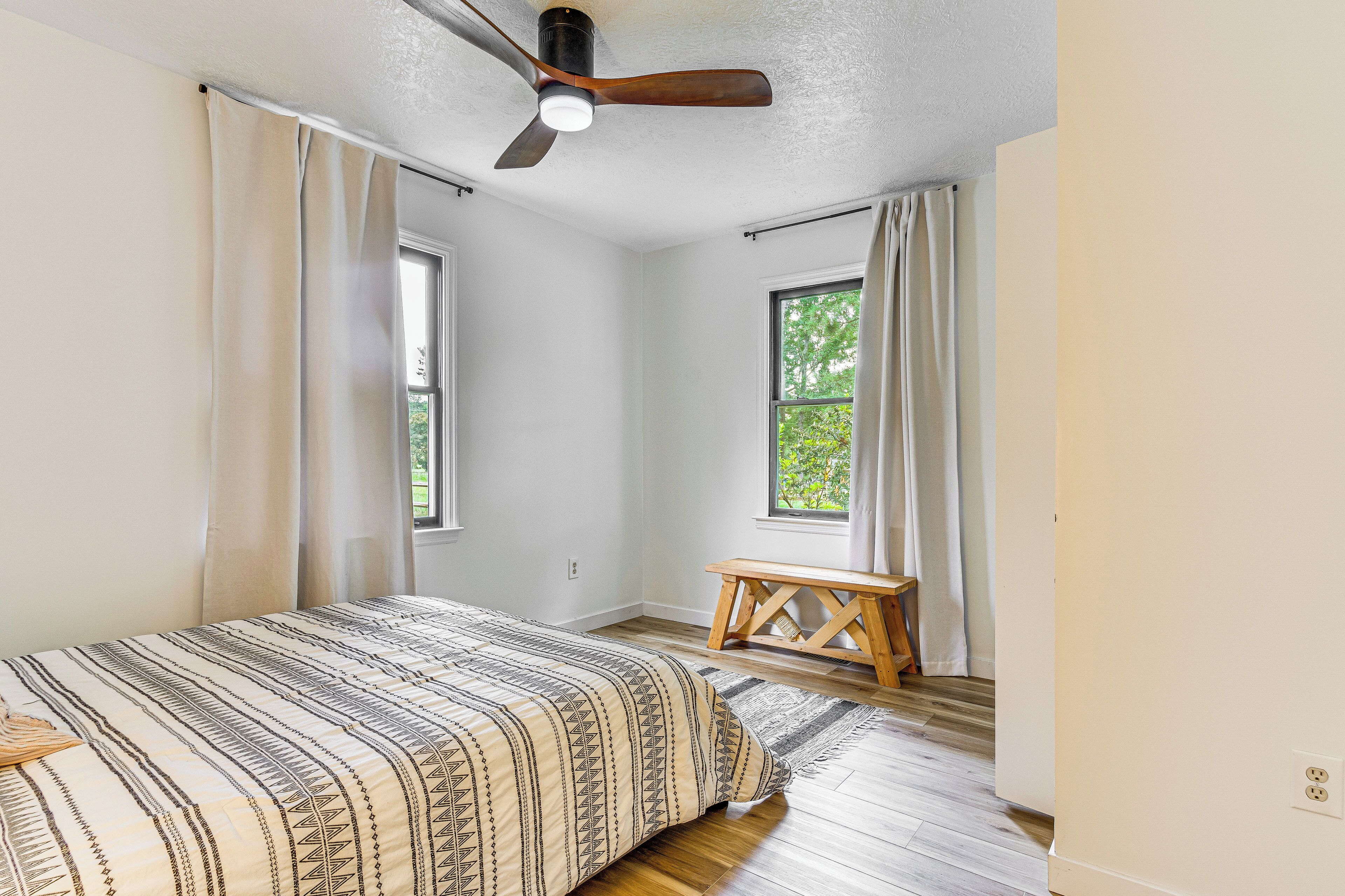 A bedroom with a bed, a ceiling fan, two windows, and a wooden bench.