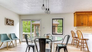 A dining area with a glass table surrounded by chairs, a kitchen with wooden cabinets, and a view of a wooden structure outside. - Private Island Getaway in Roanoke Rapids w/ Grill! (Roanoke Rapids)