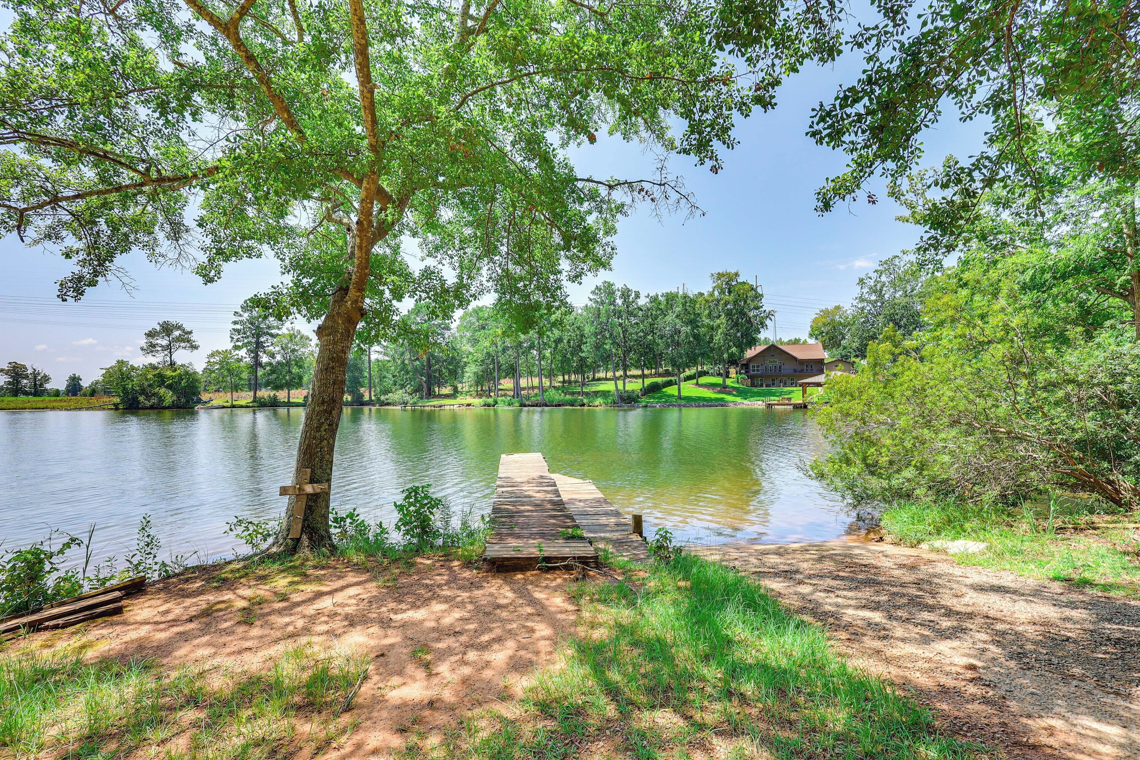 A lakeside wooden dock extending into a calm lake, surrounded by lush greenery and a small house in the background.