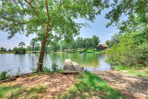 A lakeside wooden dock extending into a calm lake, surrounded by lush greenery and a small house in the background. - Pet-friendly Sparta Retreat: Porches & Fire Pit! (Milledgeville)