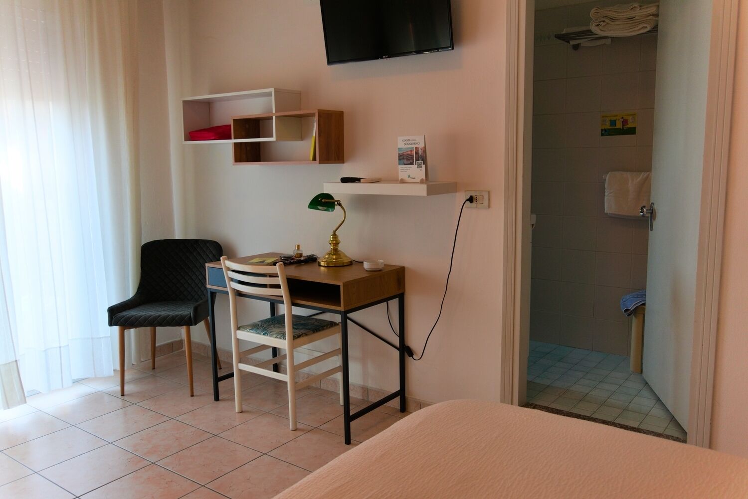 A hotel room with a bed, a desk with a lamp, a chair, a TV mounted on the wall, and a bathroom visible through an open door.
