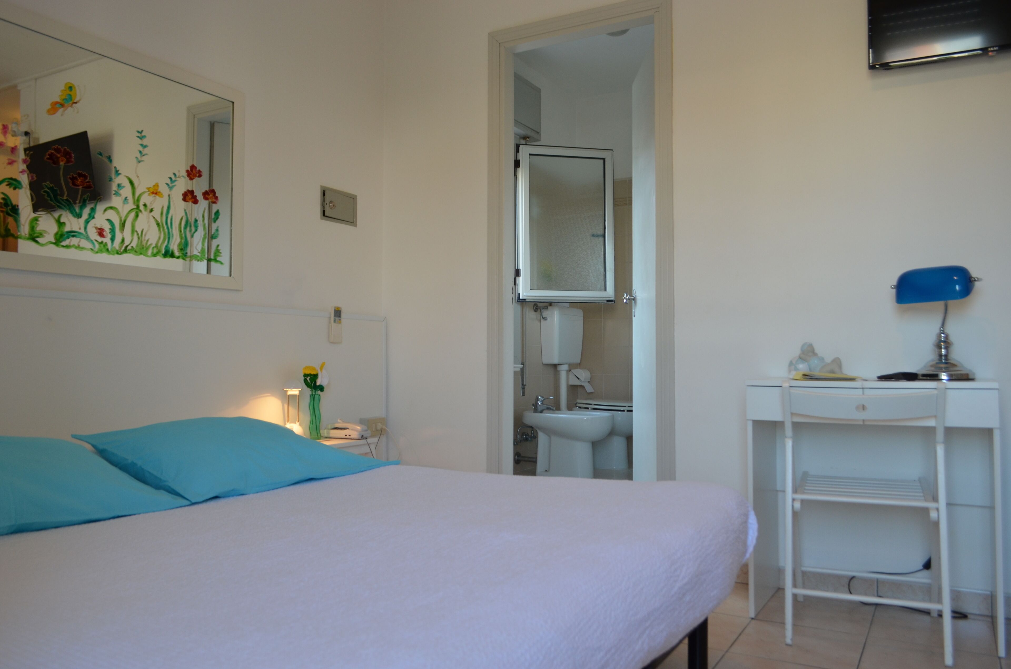 A bedroom with a bed, a bedside table, a mirror, a sink, and a desk.