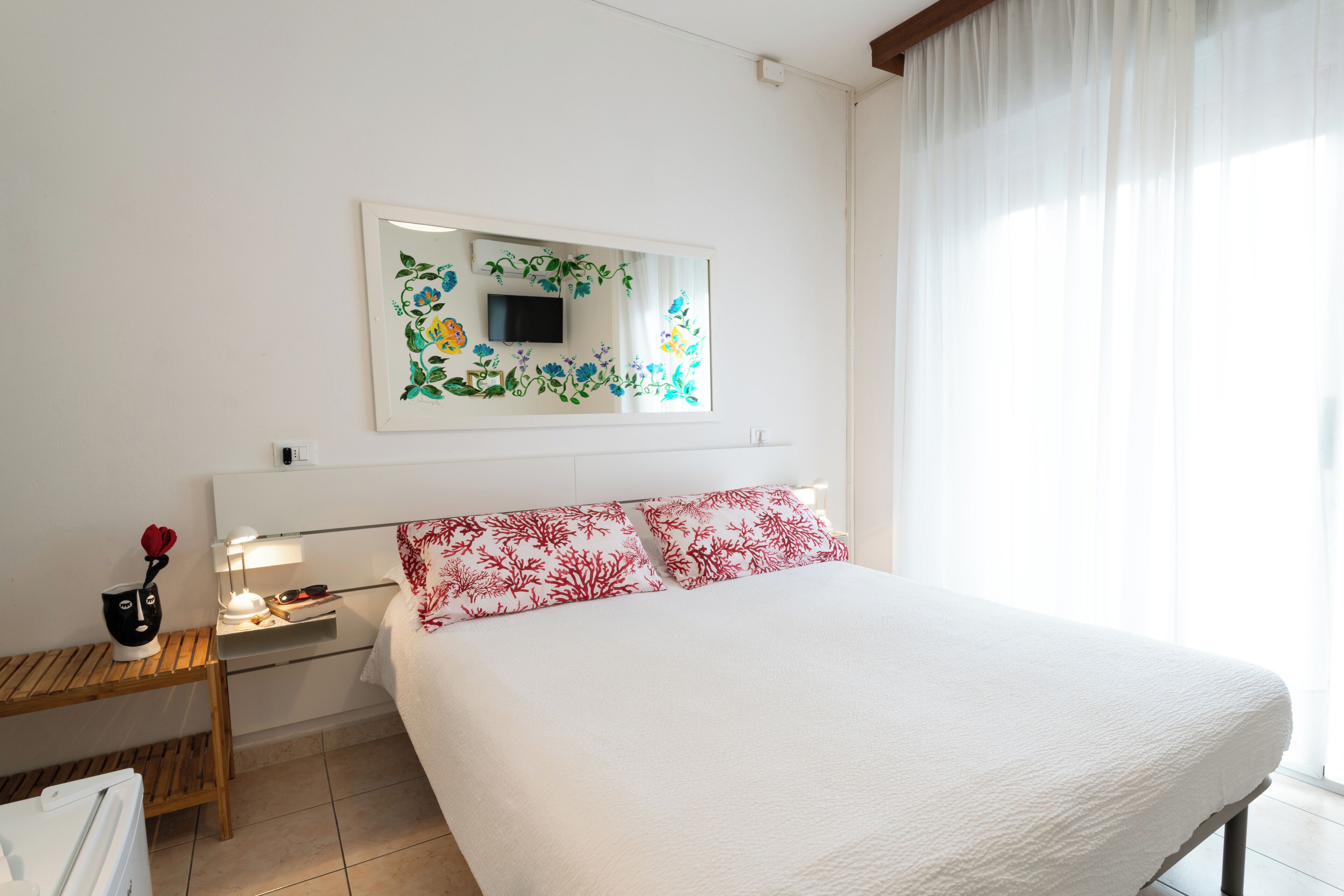A hotel room with a bed, a bedside table with a lamp, a small table with a flower vase, and a wall-mounted TV.
