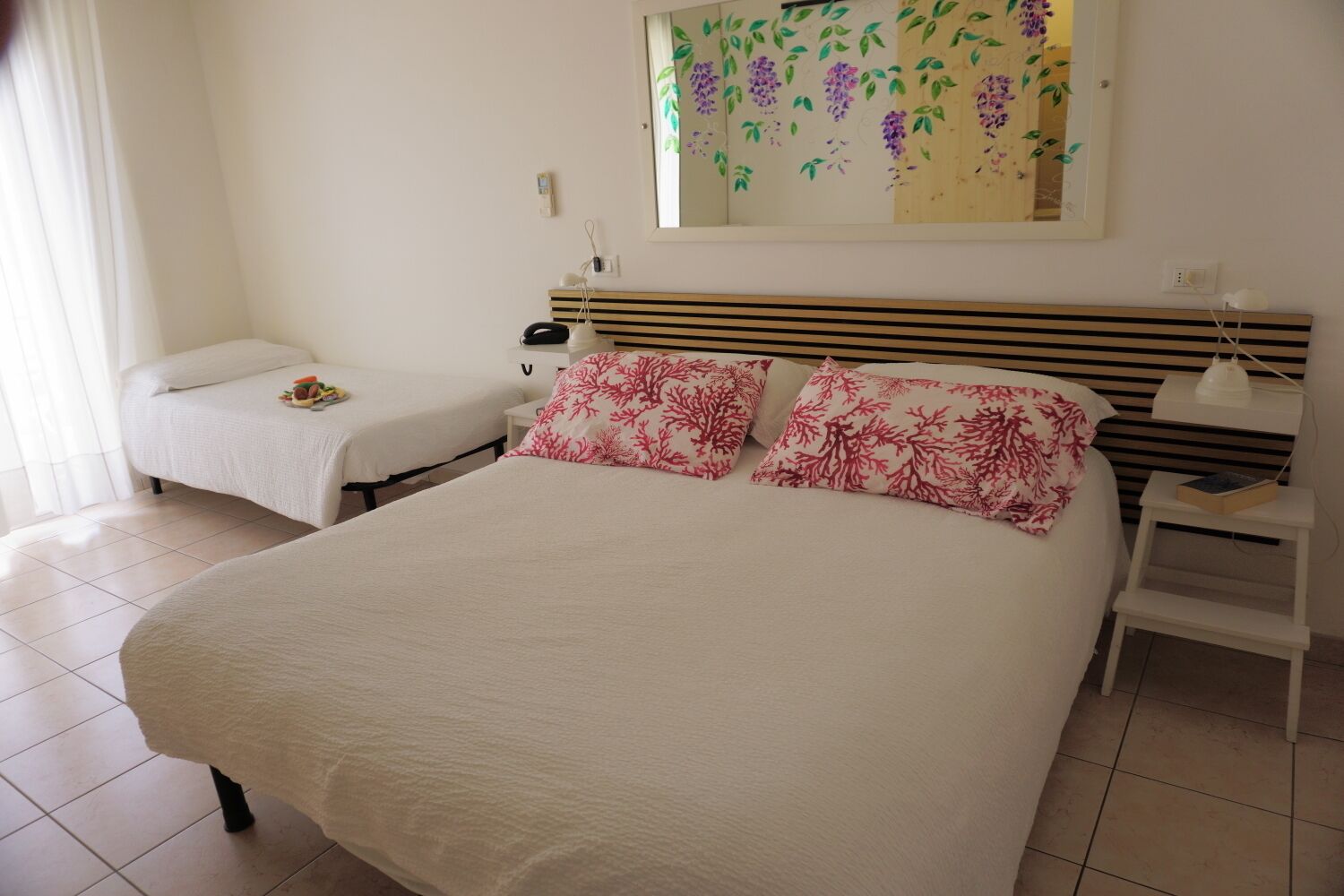 A bedroom with a large bed, two smaller beds, a nightstand, a lamp, and a wall-mounted mirror.